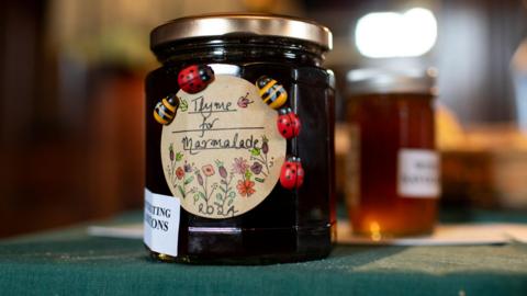 Close up view of a jar of marmalade. The hand-written label reads "Thyme for Marmalade", with pictures of flowers and models of bees and ladybirds around it. The jar is resting on a dark green cloth and out of focus behind it can be seen another jar of marmalade.