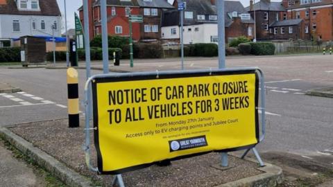 A yellow sign is cable tied onto a piece of temporary fencing, stating: "notice of car park closure to all vehicles for three weeks" with smaller text giving details of dates and electric charging points. In the backdrop are houses and hotels.