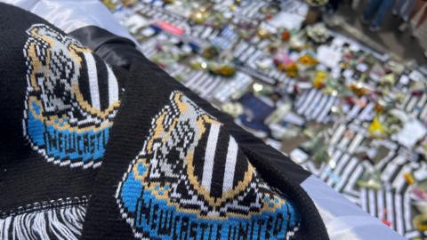 A Newcastle United scarf in the foreground. The background is blurred and shows tributes left at St James' Park for Kevin Keegan. They include team shirts and flowers.