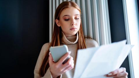 Woman standing in front of radiator looks at energy bill with phone in her other hand