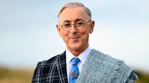 Alan Cumming smiling while wearing a blue tartan suit