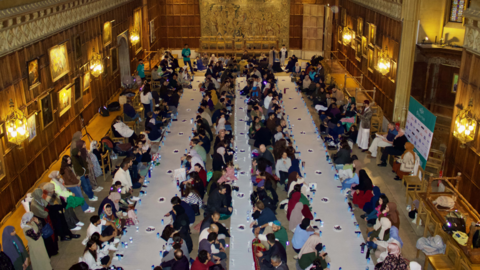 There are three rows of white cloth on the floor which span across the whole room. On either side of the rows, people are sitting lined up talking to each other. 