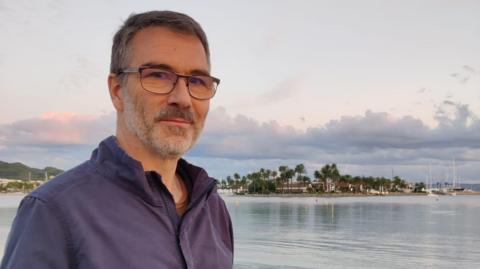 Guy Walker looking at the camera, wearing glasses and a navy shirt, with the sea behind him and a little island with palm trees and small buildings. 