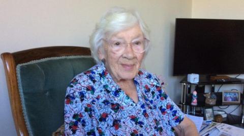 A white-haired woman wearing large plastic framed spectacles is seated in an upholstered armchair, slightly angled toward the camera. She is wearing a short-sleeved top with a floral pattern.