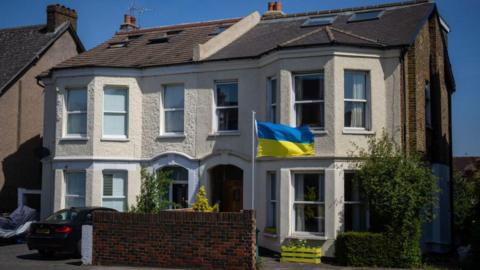 A semidetached victorian house. In the front garden a yellow and blue ukrainian flag has been erected. A window box has also been painted yellow.
