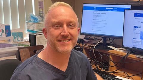 Dr Jonny Dillon is sitting at a desk. Two computer screens are behind him. He is wearing a navy uniform and has short blonde hair. Blinds and boxes cover a window directly behind him.