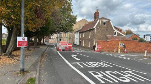 A street blocked off by signs saying no entry, with an arrow advising drivers to turn right. there are houses along the right hand side of the road