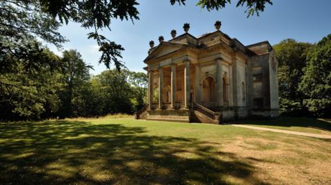 A grand, stone Greek-style building with four pillars at the front. There are large archways at the front and two staircases on either side of the entrance. On top of the roof are five stone sculptures which look like large urns. There is a patch of grass in front of the building and trees surrounding it. The sun is casting shadows of the trees on the grass and the sky is bright blue.