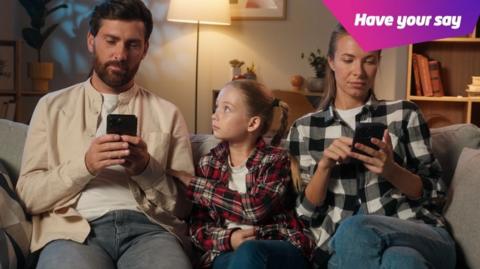 Girl sat on sofa between her parents who are both on their phones, trying to get the attention of her father.