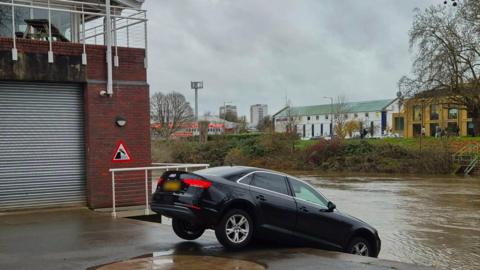 A black Audi car balances on the top step which leads down to the river. The rear wheels are off the ground and there is a sign next to the car warning drivers to avoid the drop.