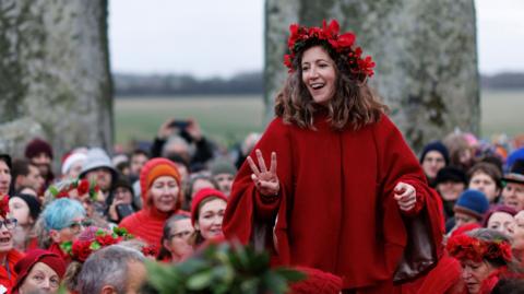 People celebrating the winter solstice at Stonehenge. A woman in a red cape and red floral headdress stands above the crowd