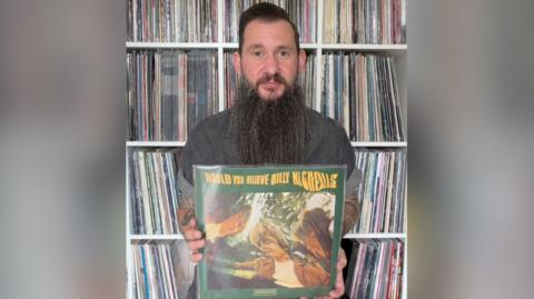 Martin is holding Billy Nicholls record, would you believe. The cover is green with yellow text. The image is a wavy effect photo of the main artists involved. Martin has dark hair and a very long beard. He is sat infant of loads of vinyl records in shelves