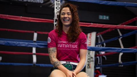 A woman with long brown hair and wearing a pink T-shirt and patterned shorts is smiling while sitting on the outside of a boxing ring in a gym.