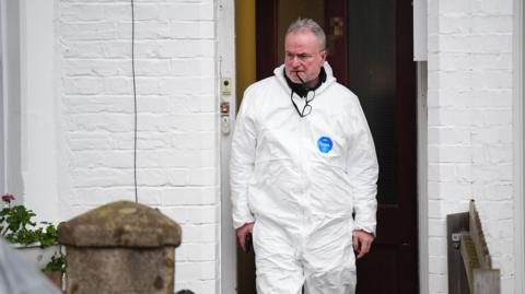 A police officer leaving the home, a white-painted terrace with a brown wood front door and geraniums in a window box. He is wearing a white all-in-one boiler suit and is chewing on the arm of his glasses while looking thoughtfully grim.