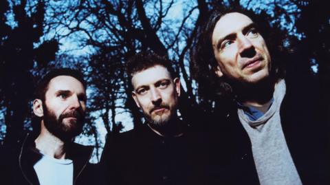 Snow Patrol band members Johnny McDaid, Nathan Connolly and Johnny Gary Lightbody. The three men appear to be in a forest, they are wearing black coats. Conolly is looking at the camera whilst the other two are looking off to the right of the frame.