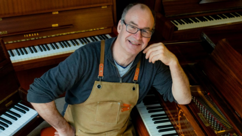The picture shows a man wearing a heavy canvas workshop apron, sitting among several upright and grand pianos inside what appears to be a restoration or tuning workshop. The person is resting one arm on the edge of an open piano where the strings and hammers are visible, while other pianos surround the scene, their lids raised to show keyboards and wooden casings.
