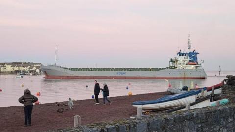 A large silver cargo ship called Scot Pioneer is seen in the water in Teignmouth Harbour. Three adults a small child are seen in the foreground on the beach at Shaldon.