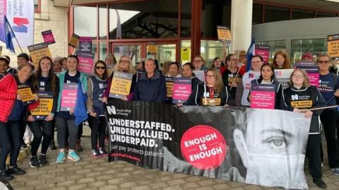 A group of nurses holding RCN placards and a banner with the words 'Understaffed, Undervalued, Underpaid' and 'Enough is Enough' on it.