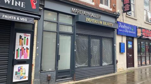 A salon in a row of shops. The building is grey with a door in the middle. Above it, in gold lettering, it says "Imperium Beauty".