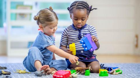 Stock image of two toddlers - one girl and one boy - playing with toy bricks on a carpet