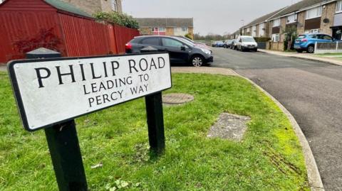 An estate made up of light brown terrace houses. In the foreground on a patch of grass is a sign which says Philip Road, leading to Percy Way