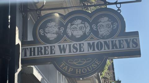 A close up of the Three Wise Monkey pub's sign outside the pub. Three monkey heads are on the sign each with a different expression. 