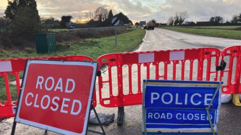 Road closure signs in front of red barriers that have been put up on Cirencester Road. Emergency service vehicles can be seen in the distance.