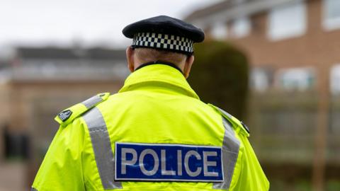Generic image of a police officer wearing a black cap with a white and black band on it. They have their back to the camera and are wearing a yellow jacked with a blue sign on the back with the word police written on it 