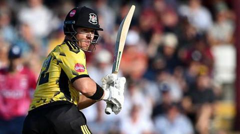 D'Arcy Short, wearing a black helmet, yellow shirt, black trousers, black pads and white gloves, plays a cricket shot while holding his bat