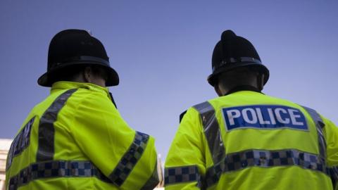 Stock image taken from the back of two police officers stood side by side wearing fluorescent police jackets and black police hats.