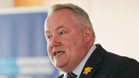 A man with white hair, Darren Millar, is wearing a navy suit and pointing while making a speech at a lectern.