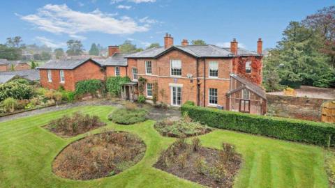 A brick-built Georgian seven-bedroomed house in Oswestry with hedges, a lawn and flower beds in the foreground.