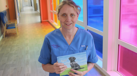 Nurse Lin Fidgin stands in a hospital ward with brightly coloured windows and holds up a children's book which has a title called Bess of Clara Vale. She has blonde hair which is tied back and is wearing blue nurse scrubs.