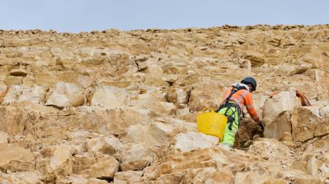 An engineer wearing hi-vis work gear and a helmet is connected to a rope as he works on a limestone escarpment in the Cotswolds