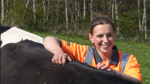 Claire is standing next to a cow and rests her arm on the animal. She has an orange top on and brown hair tied back. She is smiling at the camera in the sunshine.