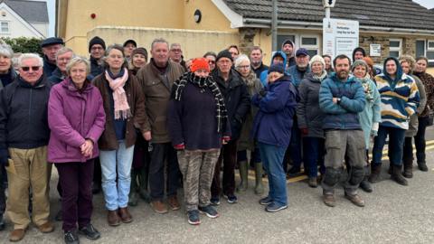 About 30 people standing in a car park front of the public toilets at Porthcothan Bay. They are bundled up in coats, scarfs and hats.