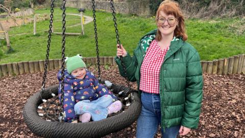 A picture of Freya (left) in a swing and her mum Amy (right) in their local park. Freya is smiling and wearing a coat and jeans. Amy has ginger hair and glasses and is also smiling a wearing a coat and jeans. There is grass and playground equipment in the background.