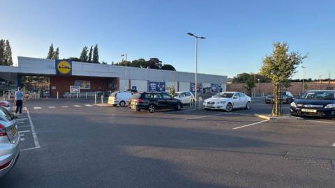 A Google image street view showing the Lidl car park in Stroud. It is nearly dusk but the sky is still a bright blue. There are eight cars parked near the entrance. 