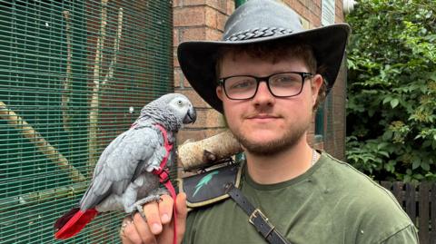 A picure of Connor Atherton who is wearing a hat and green t-shirt and black glasses. He smiles into the camera, whilst holding a gray parrot.