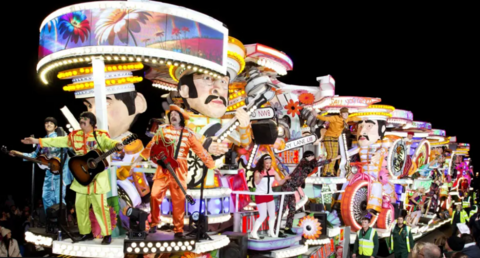 A carnival cart with people dressed as The Beatles and in 1960s fashions. It is brightly lit and decorated with heads of The Beatles, London street signs and flowers. There are people wearing hi-vis jackets walking alongside.