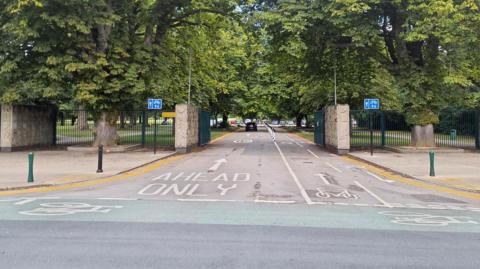 A road entrance to a park with large open gates, bollards, road markings and trees.