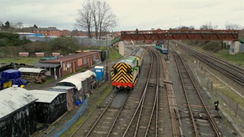 A green diesel shunter on a railway line heading under a footbridge. The engine has black and yellow diagonal stripes on the back and a red bumper. Alongside the track are a collection of sheds