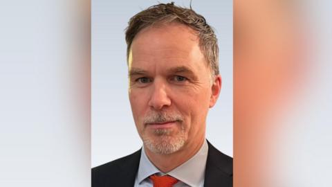 Stephen Wright has short brown hair which is greying at the sides. He has a neat goatee beard that is mostly grey. He is staring at the camera and is wearing a dark suit jacket with a light shirt and red tie.