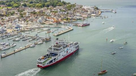 A stock image of a Red Funnel ferry in the sea, near a port with houses and other boats around the ferry and by the port too. 