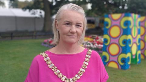 Lord Mayor Tracy Kelly is wearing pink and a gold chain of office. She has short blond hair. She's standing with grass and trees behind her. Some colourful stands have been erected.
