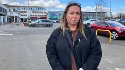 A woman with brown hair in a busy car park and lots of shops in the background.