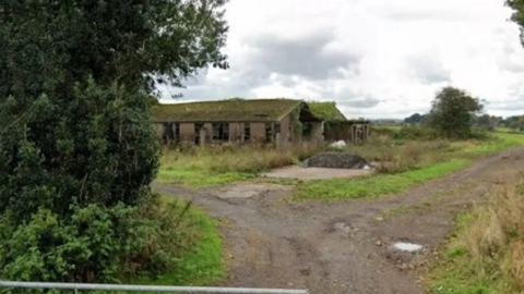 A single-storey building with its windows and doors missing and a green-covered roof at the end of a footpath with bushes in the foreground