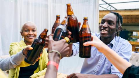 Casually dressed men and women in their 20s sitting together and smiling as they lift beer bottles for celebratory toast.