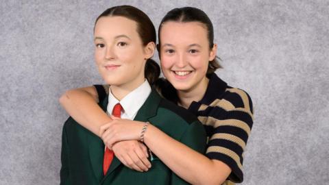 Bella Ramsey wearing a black and cream hooped top stood behind a waxwork of them, with their arms around the waxwork's shoulders.