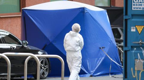 A blue forensic tent is erected in the street with an officer dressed in white overalls walking towards to. A black car is parked next to the tent